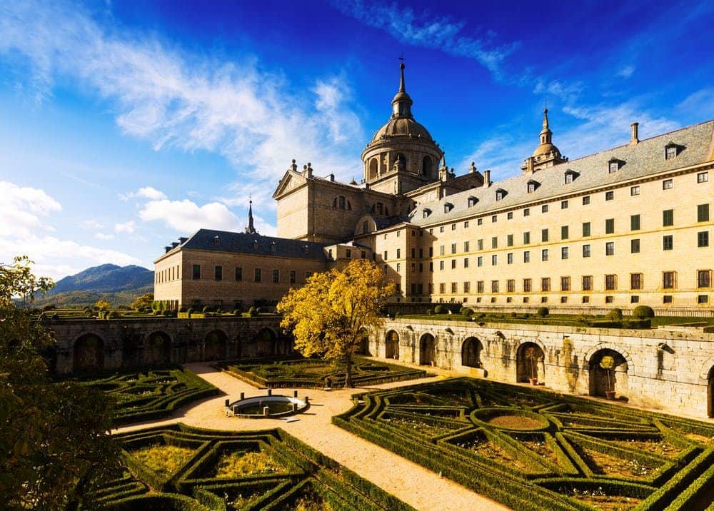 Royal Monastery of El Escorial + Valley of the Fallen: Half Day Tour from Madrid A historic stone building with multiple spires and a domed tower behind an ornate garden with hedges and a central tree.