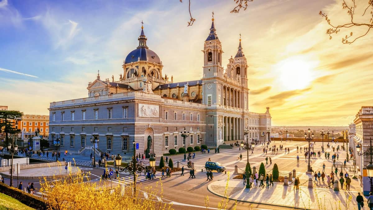 A grand building with domes and towers under a sunset sky, surrounded by people, cars, and manicured gardens.