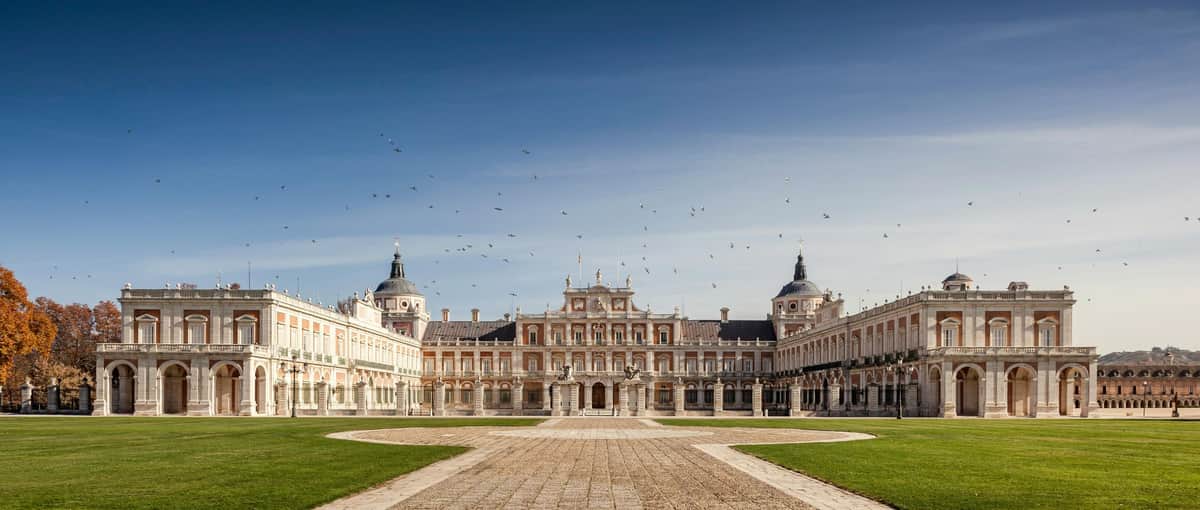 Royal Palace of Aranjuez: Entry Ticket + Digital Royal Guide A grand palace with two domed towers, a central building, and a wide courtyard, under a clear sky with scattered birds.