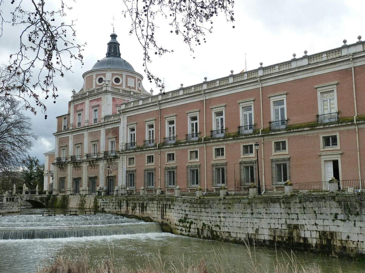 Royal Palace of Aranjuez: Guided Tour Historic building with a dome, pinkish brick facade, and multiple windows, situated next to a cascading river under a cloudy sky.