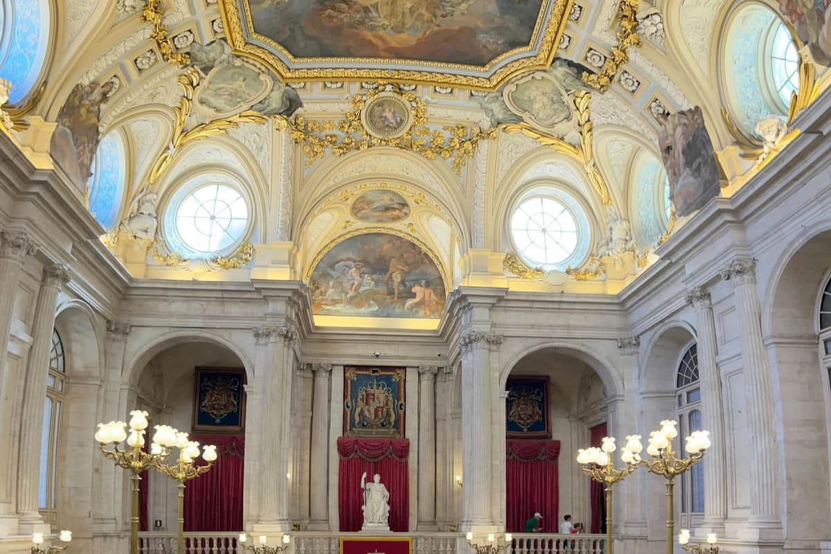 An ornate hall with a decorated ceiling, round windows, red drapery, a statue, and chandeliers. People are visible in the scene.