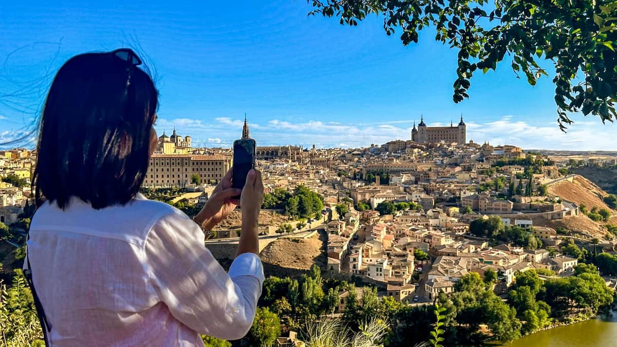A person photographs a historic city with numerous beige buildings and two prominent towers under a clear blue sky.