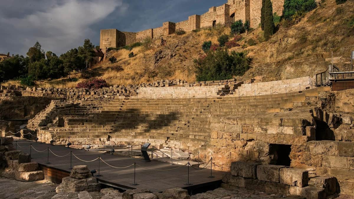 Málaga Roman Theatre & Alcazaba: Guided Tour Ancient stone amphitheater with tiered seating, surrounded by ruins and vegetation under a partly cloudy sky.