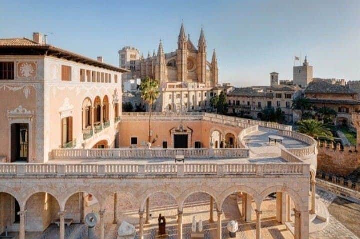 A sunny view of a historic cathedral and surrounding buildings, featuring arches and a courtyard in the foreground.
