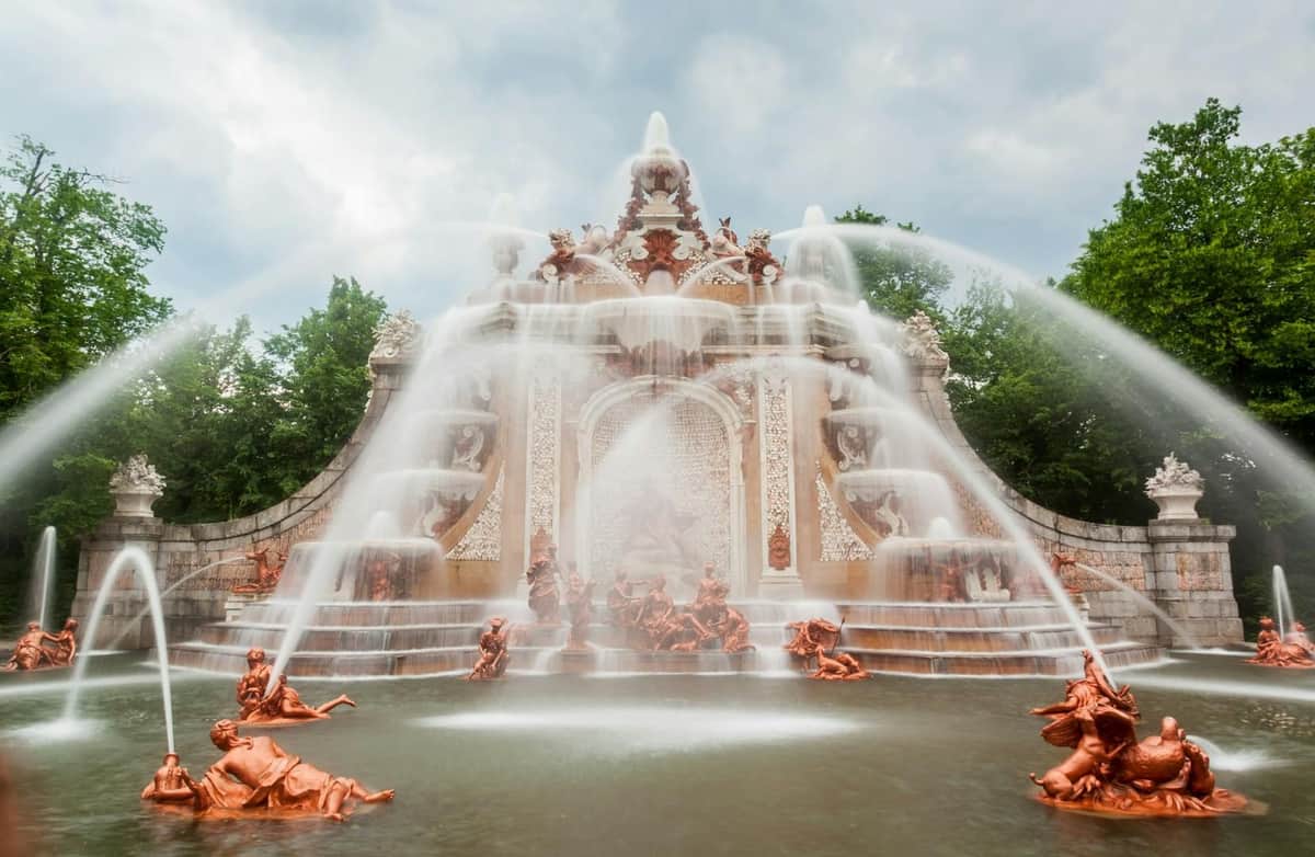 An ornate fountain with multiple tiers and sculptures, spraying water elegantly, surrounded by trees under a cloudy sky.