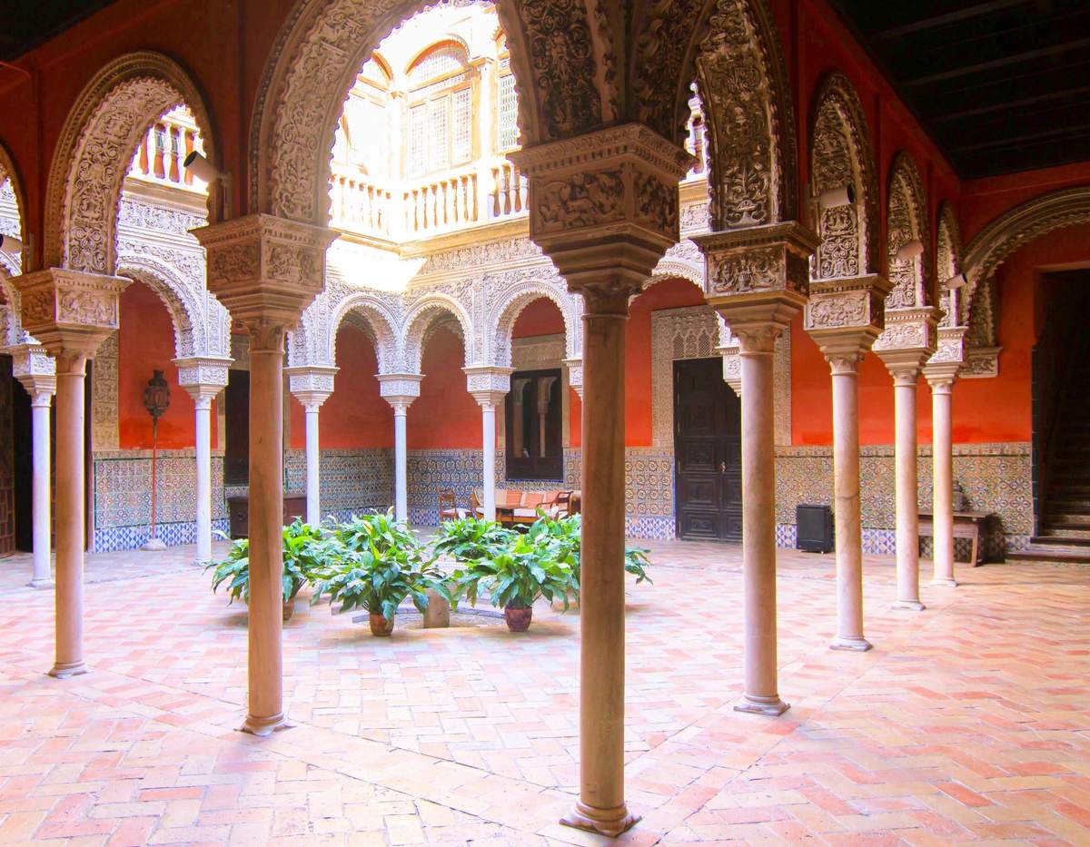 A courtyard with ornate arches, potted plants on tiled flooring, and decorated walls under a bright natural light.
