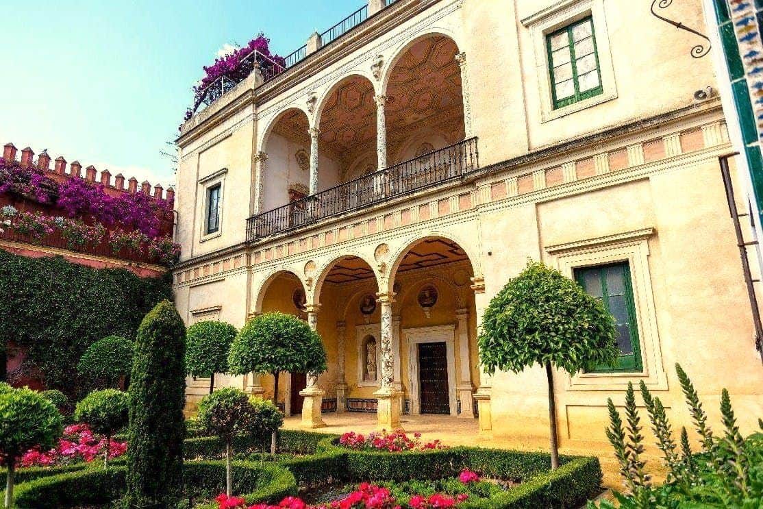 Palacio de las Dueñas y Casa de Pilatos: Tour guiado Edificio ornamentado de dos plantas con balcones arqueados, jardín cuidado y flores de colores en primer plano.