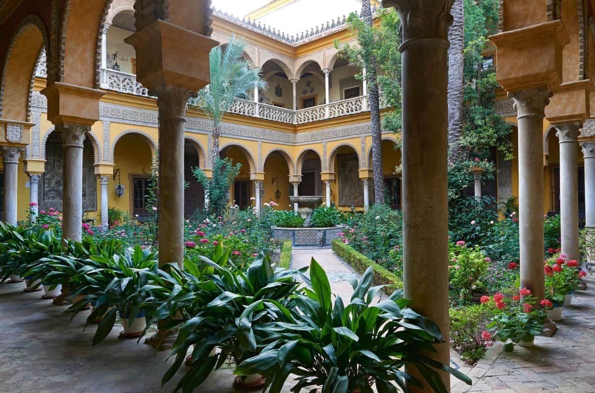 A courtyard with lush greenery, potted plants, arched yellow buildings, and a central fountain surrounded by flowers.