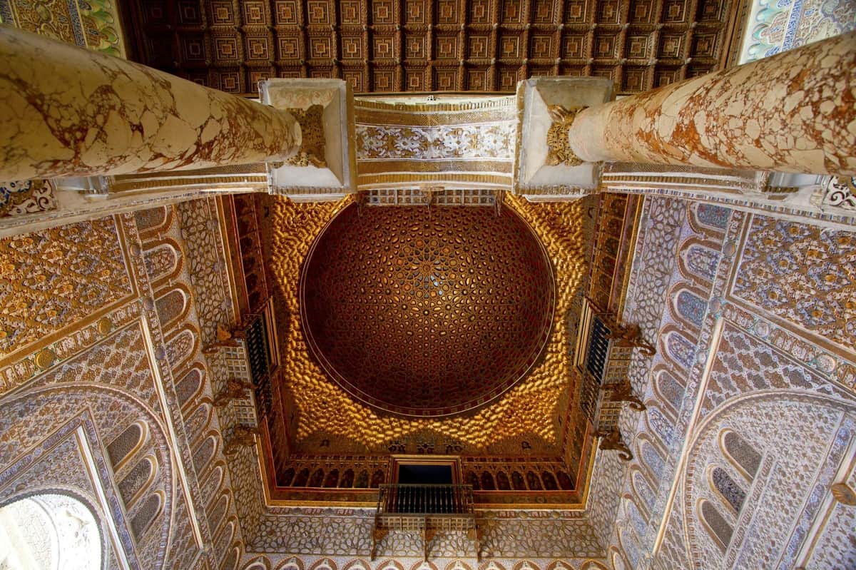 Royal Alcázar of Seville & Seville Cathedral: Small Group Guided Tour A richly decorated ceiling with intricate patterns and a golden dome, surrounded by ornate walls and columns.