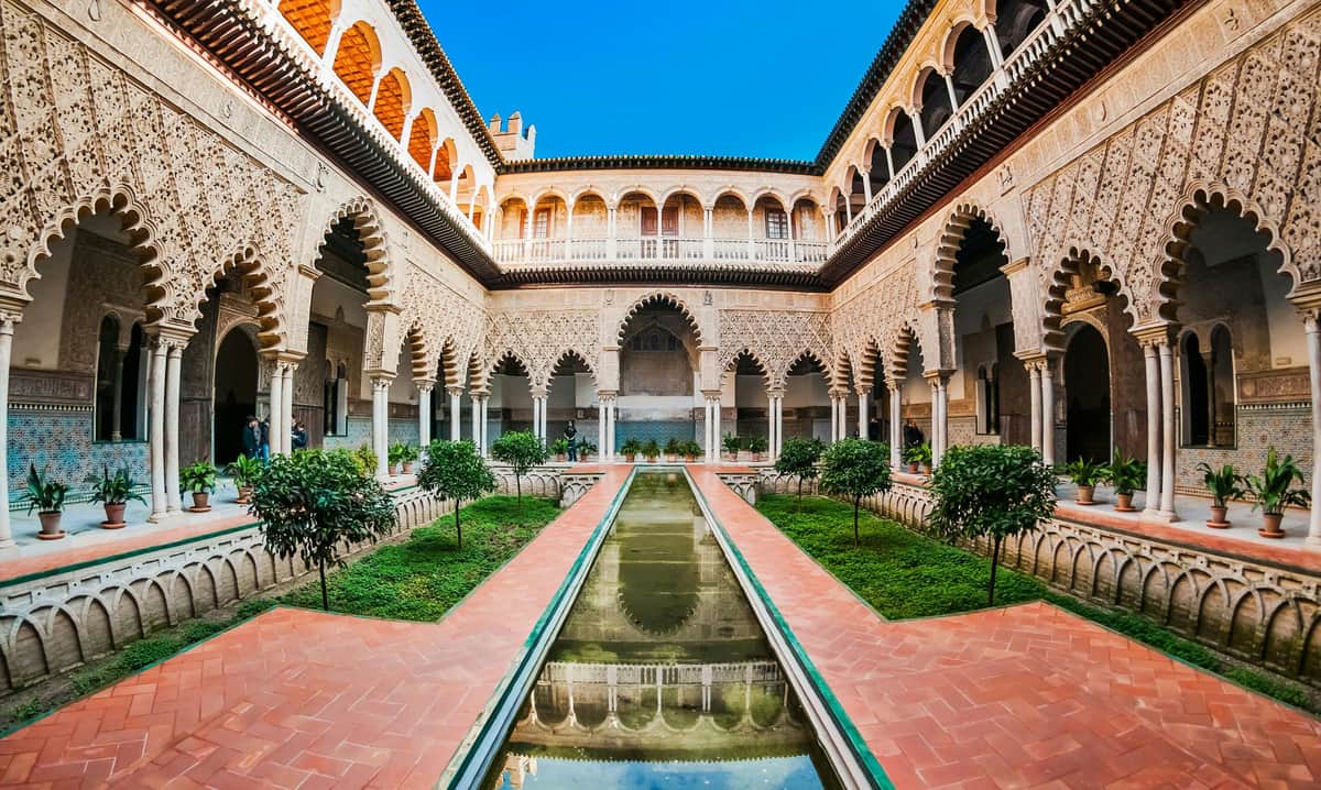 A courtyard with arched columns, a rectangular reflecting pool, and greenery, enclosed by a two-story ornate building.