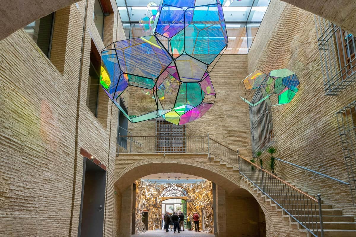 A modern atrium with brick walls, colorful geometric hanging sculptures, and people walking through an ornate archway.