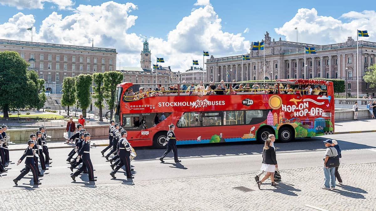 A red double-decker tour bus labeled "Stockholm Red Buses" with tourists on top, passes by a marching band and historic buildings.