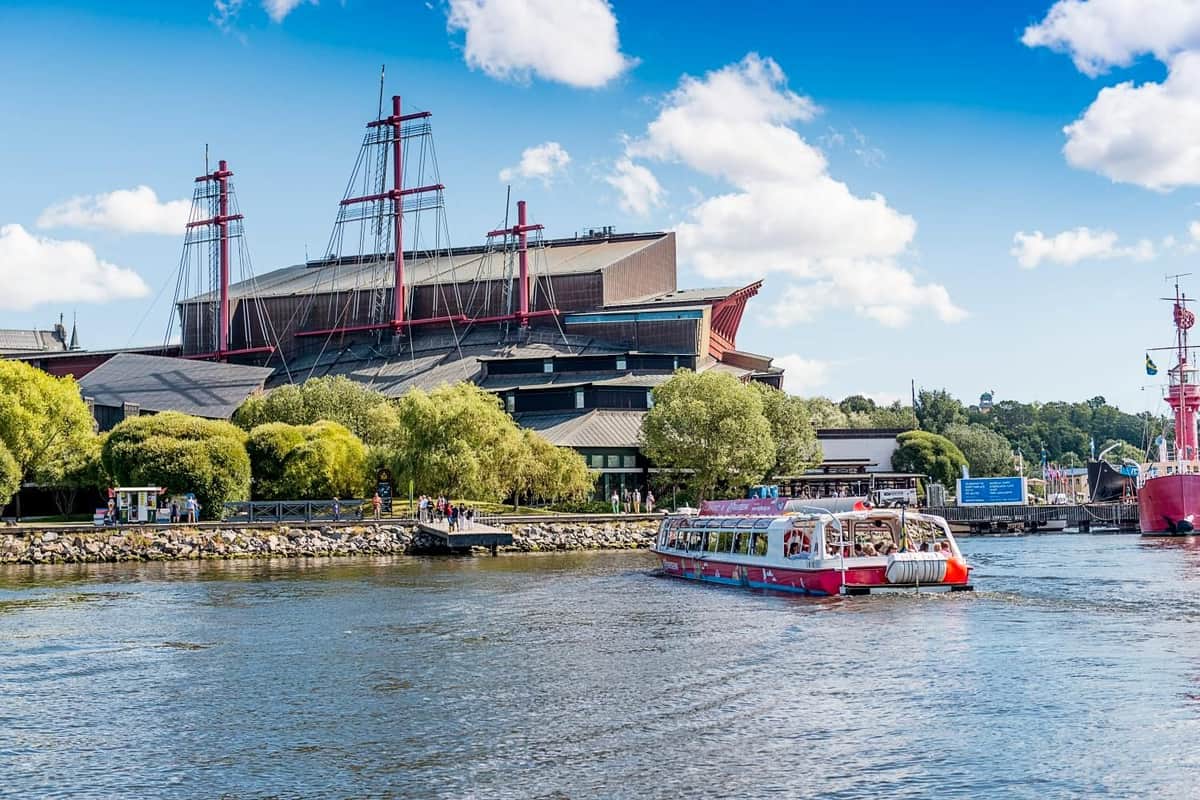A red and white tour boat on a river, with a large building and lush trees along the riverbank under a blue, partly cloudy sky.