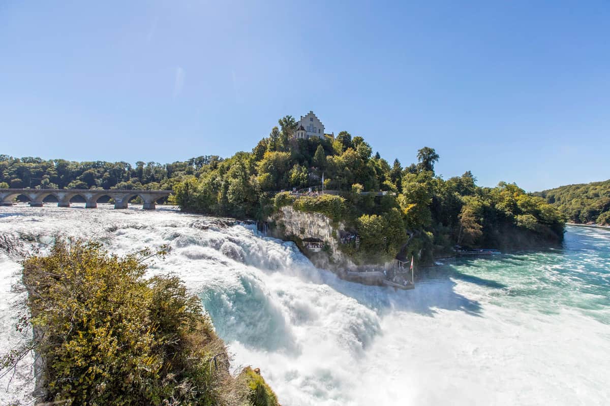 Zurich Highlights A large waterfall surrounded by lush greenery and trees with a castle-like building on a hilltop under a clear blue sky.
