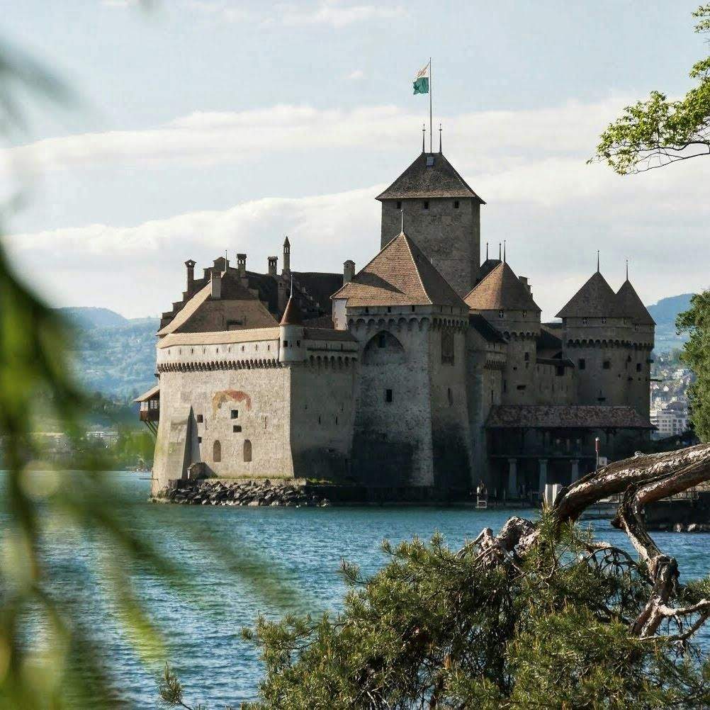 Medieval castle with pointed towers on a lake, surrounded by greenery, under a partly cloudy sky.