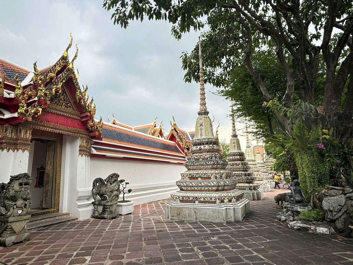 Ornate temple complex with decorative spires, red and gold roofed buildings, and stone statues, surrounded by trees and paved paths.