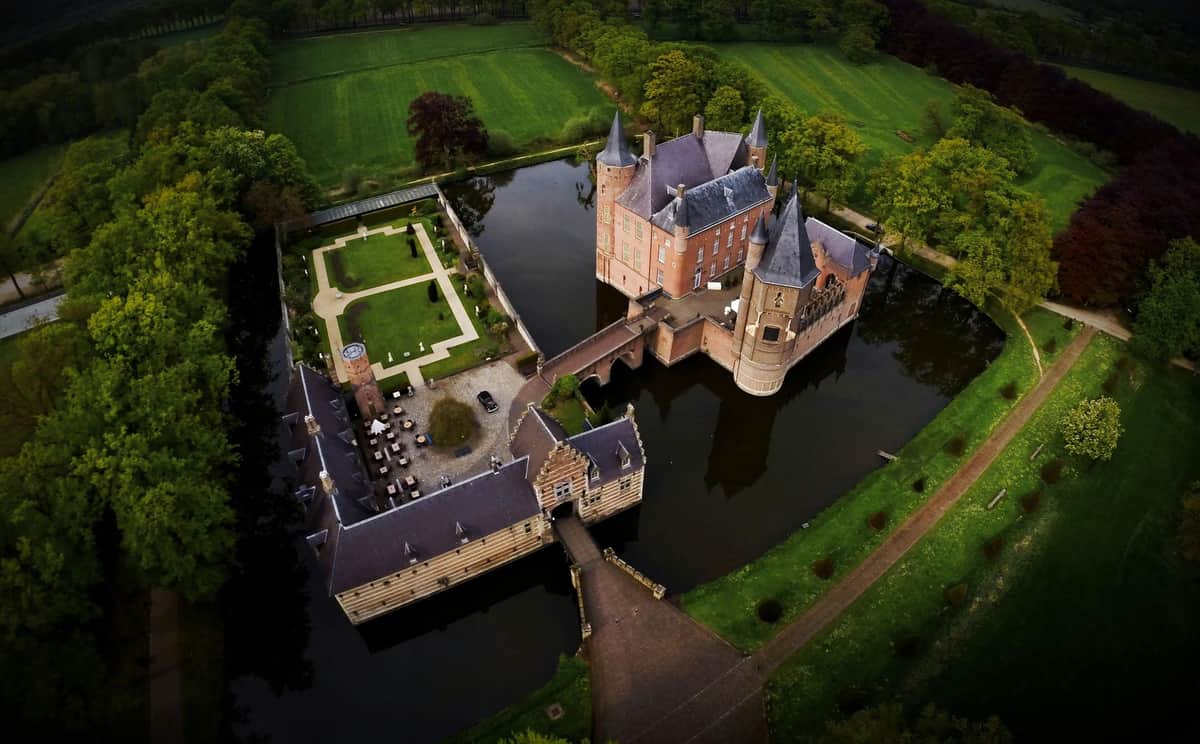 Aerial view of a castle surrounded by a moat, adjacent courtyard, and gardens with lush greenery in the background.