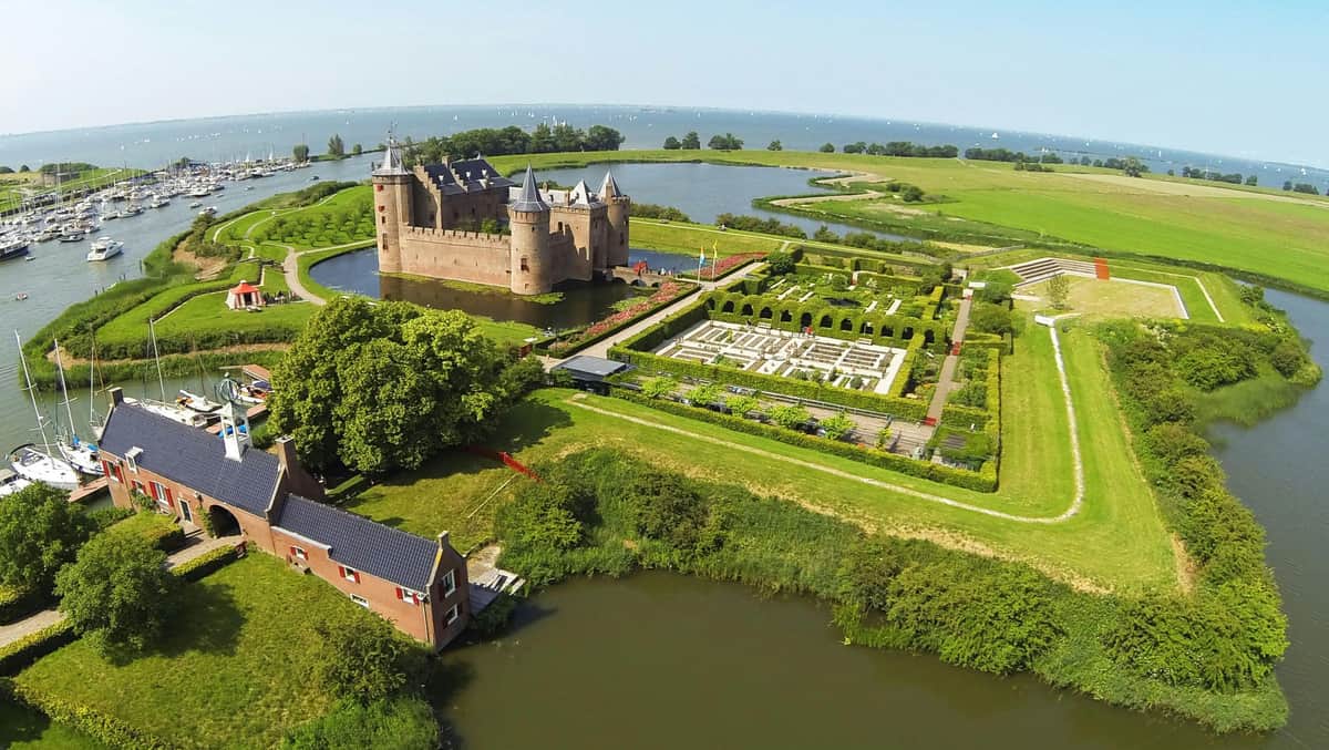 Aerial view of a castle with a moat, adjacent gardens, a body of water in the background, and a nearby smaller building.