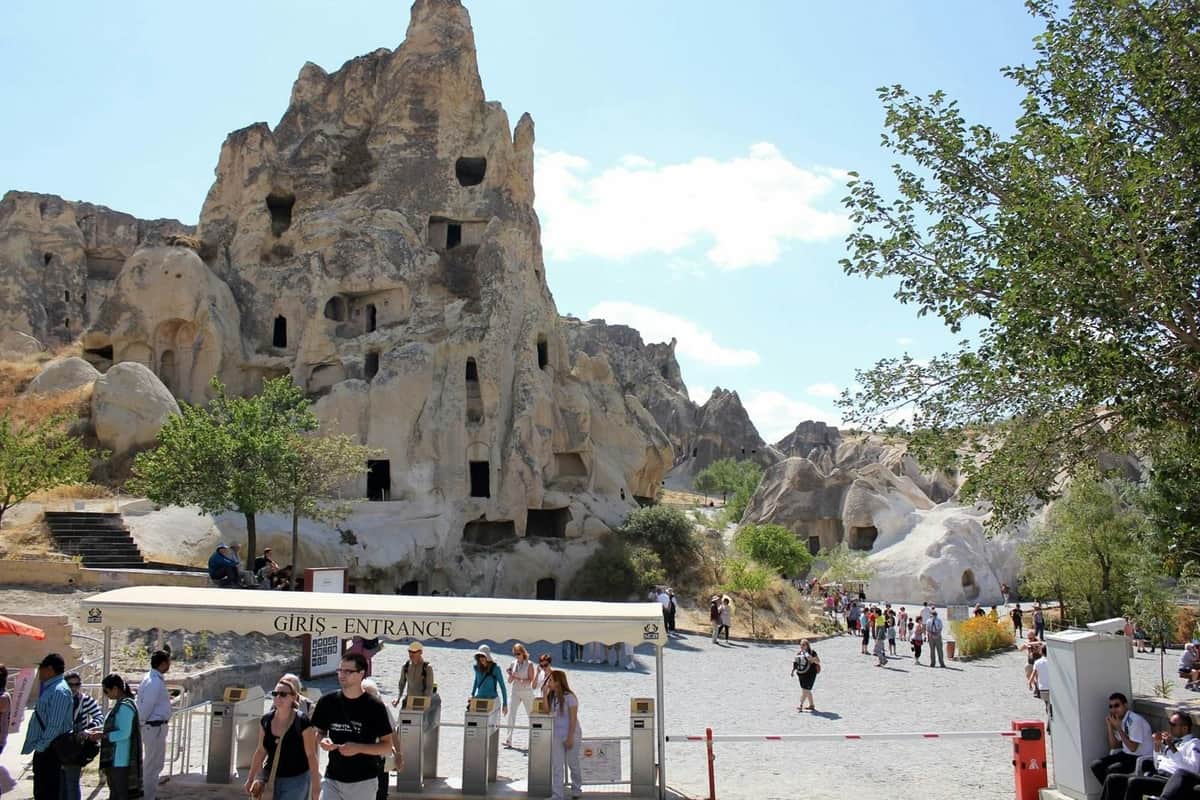 Tourists at the entrance to a rocky cave with multiple openings in Cappadocia under a clear sky.