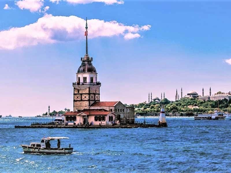 Una torre storica su una piccola isola in uno specchio d'acqua con una barca in primo piano e un paesaggio urbano sullo sfondo sotto un cielo blu.