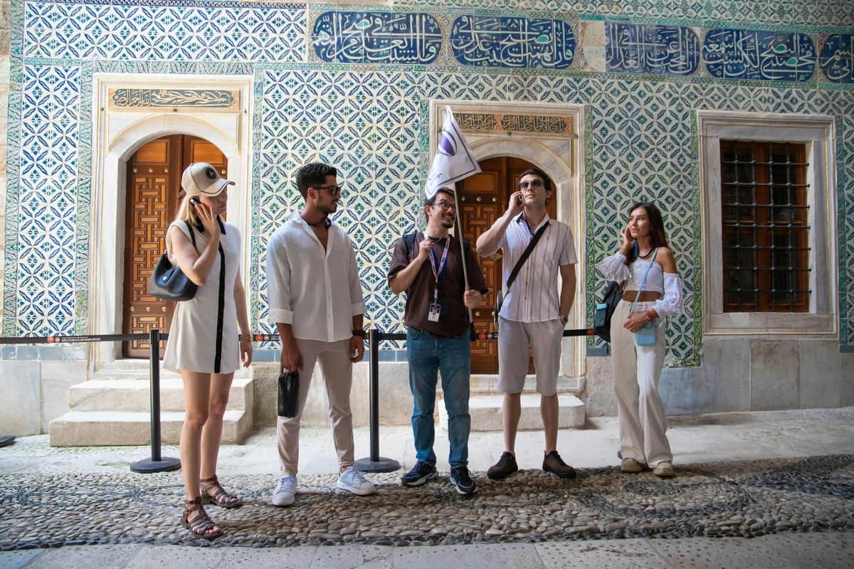 Four people stand in front of a patterned wall with Arabic script. One person holds a flag, possibly leading a tour.