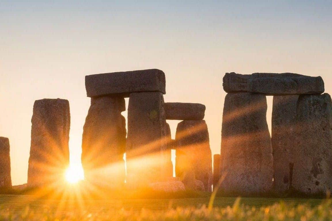 From London: Choice of Stonehenge, Windsor Castle & Roman Baths Sunlight shining through the stone structures of Stonehenge at sunset, with grass in the foreground.