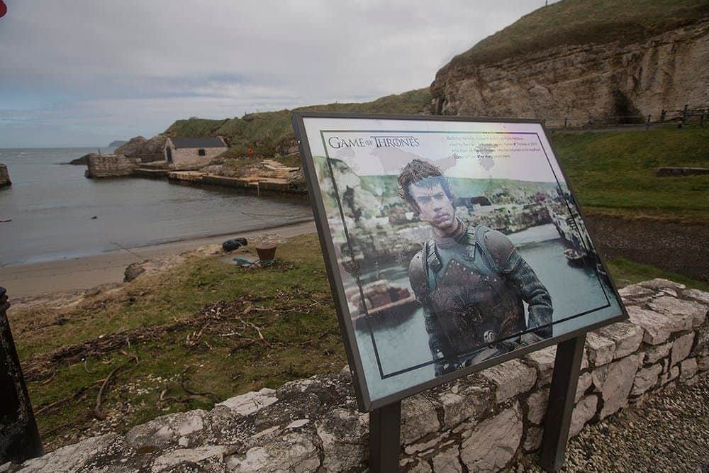 Giant’s Causeway Tour + Game of Thrones Filming Locations Tour from Belfast Informational sign featuring a character from "Game of Thrones" with a scenic backdrop of hills and water.