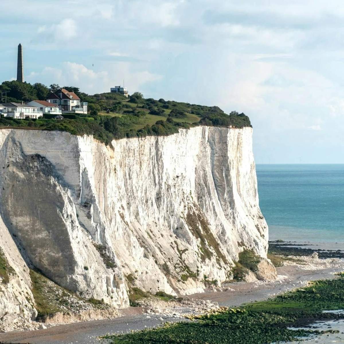 Canterbury, o Castelo de Dover e as Falésias Brancas: Visita guiada a partir de Londres Falésias brancas ao longo de uma linha costeira com casas e um monumento alto no topo, encontrando-se com o mar azul calmo sob um céu parcialmente nublado.