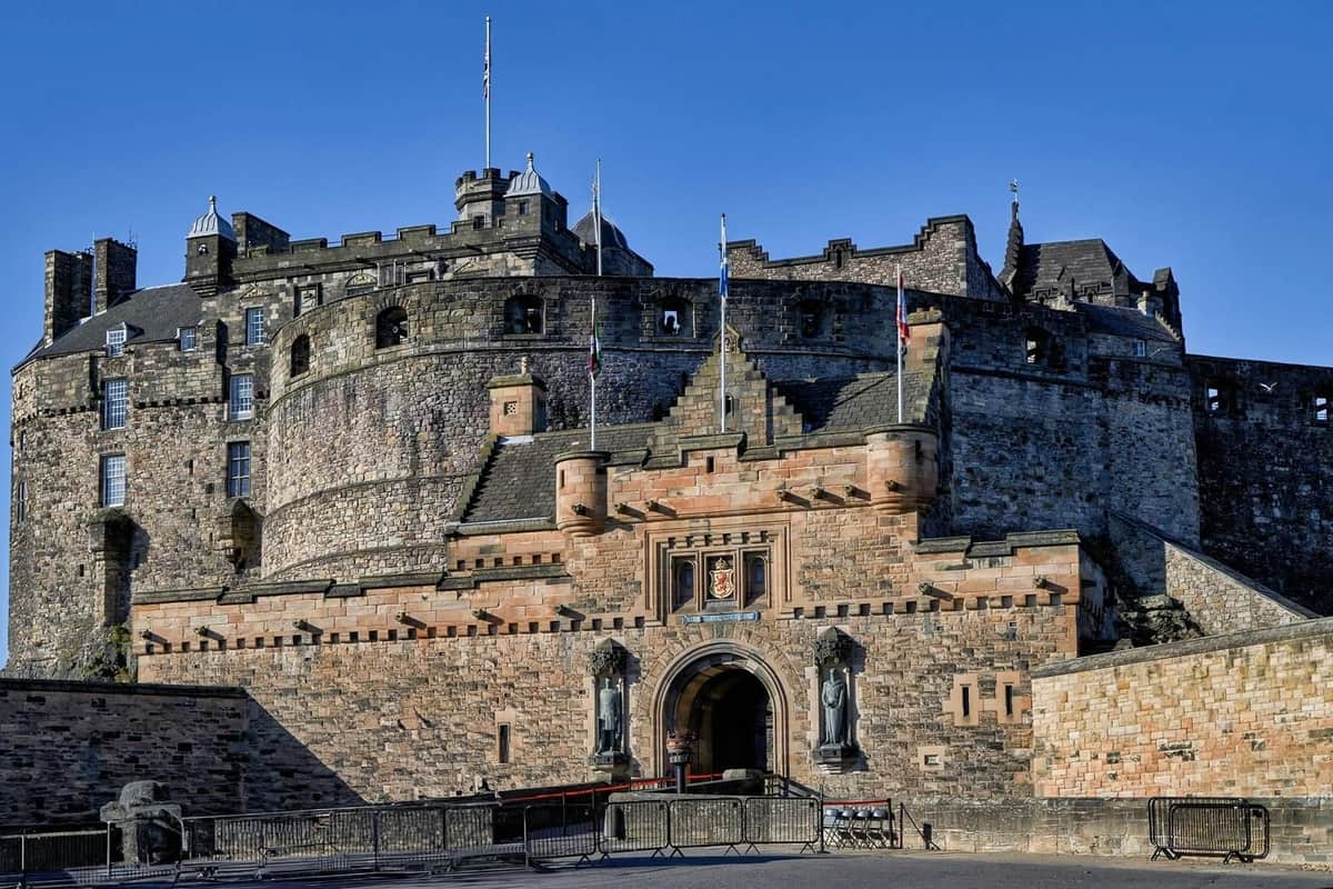 A historic stone castle with a prominent arched entrance, flags, and statues, under a clear blue sky.