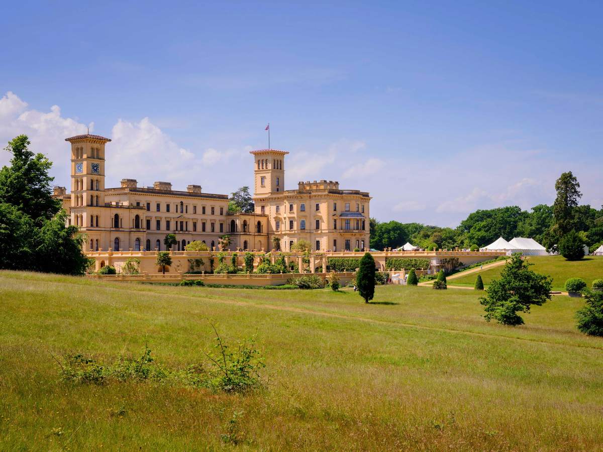 Large, historic building with towers surrounded by a green lawn and trees under a clear blue sky.