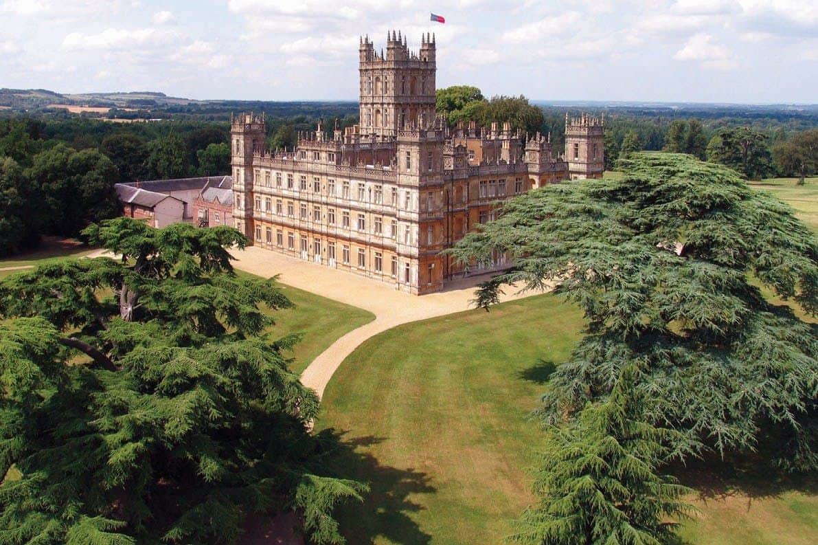 Bampton: Downton Abbey Filming Locations Guided Tour + Highclere Castle Entry A large, ornate castle with towers and a flag on top, surrounded by green lawns and trees under a partly cloudy sky.