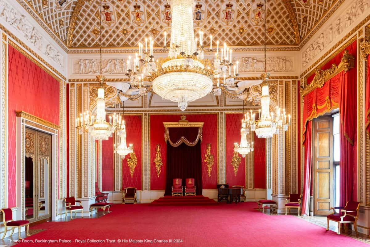 Sala opulenta com paredes vermelhas, candelabros ornamentados, detalhes dourados e duas cadeiras reais numa plataforma sob um dossel.