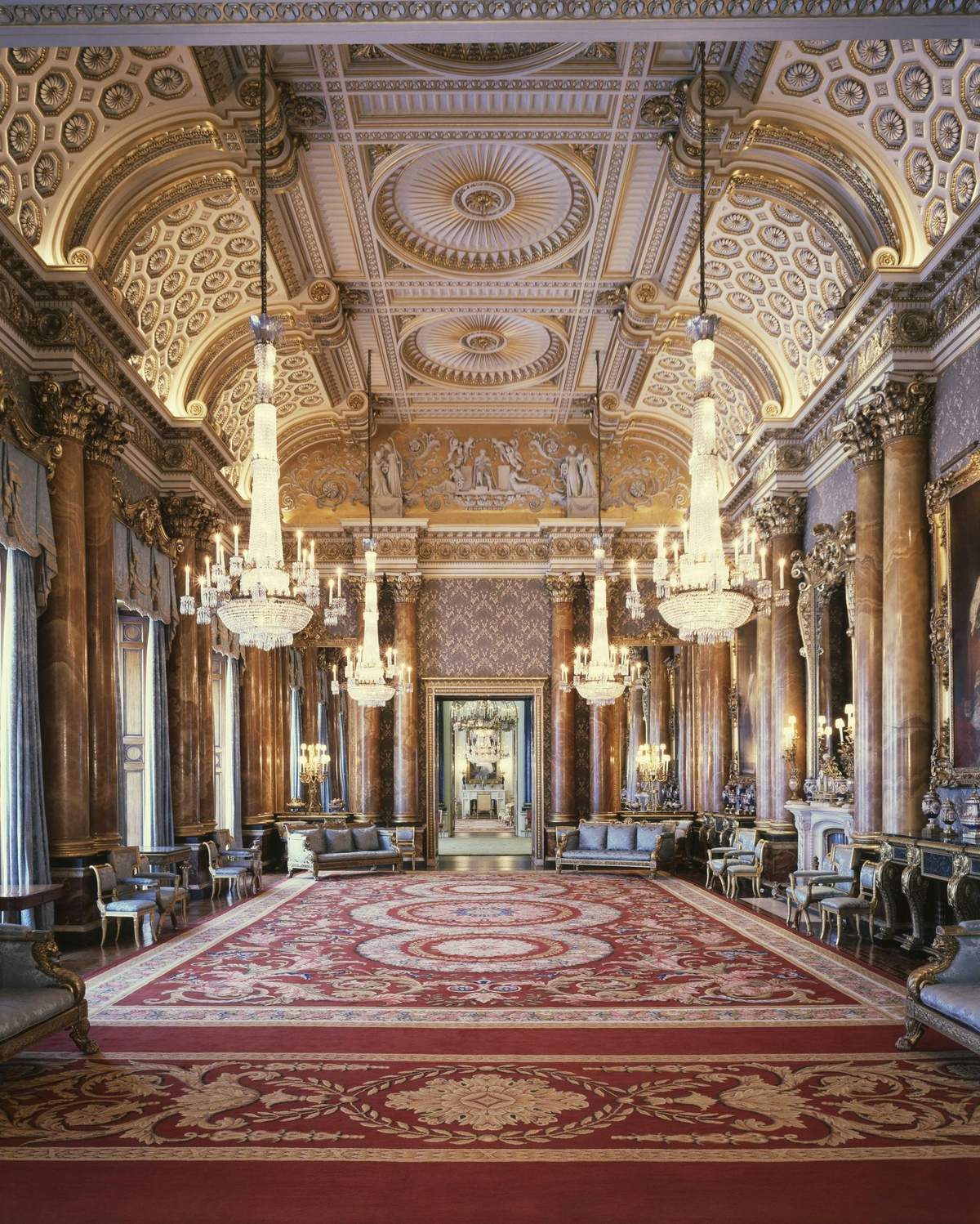 Buckingham Palace: State Rooms Admission with Audio Guide Ornate grand hall with chandeliers, detailed ceiling, marble columns, and red carpet. Furniture lines the sides, with large mirrors and paintings.