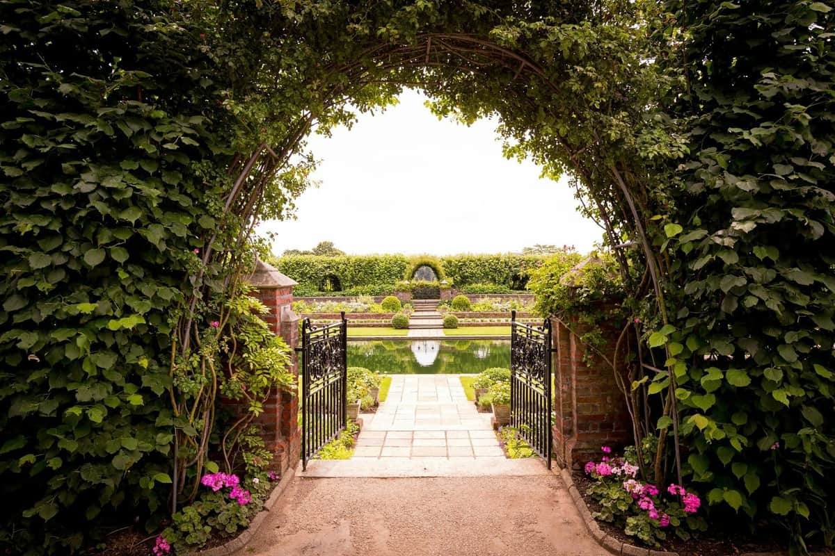 London: Guided Walking Tour + Changing of the Guard + Kensington Palace Entry Wrought iron gate opens to a brick path leading to a reflective pond, with manicured gardens, flowers, and an arched trellis.
