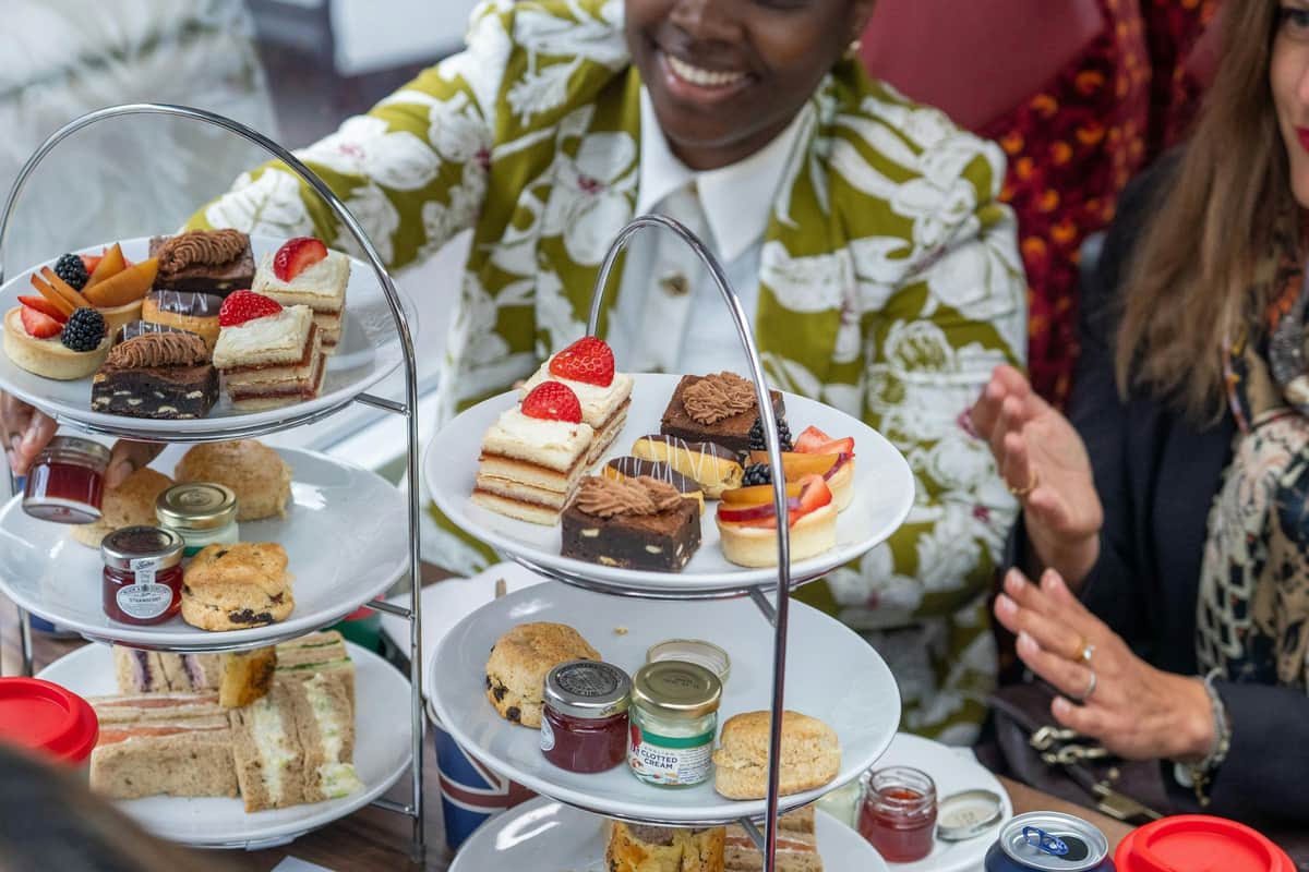 Personnes dégustant un thé de l'après-midi avec un plateau à trois niveaux composé de desserts, de scones et de sandwiches assortis. Visage souriant partiellement visible.