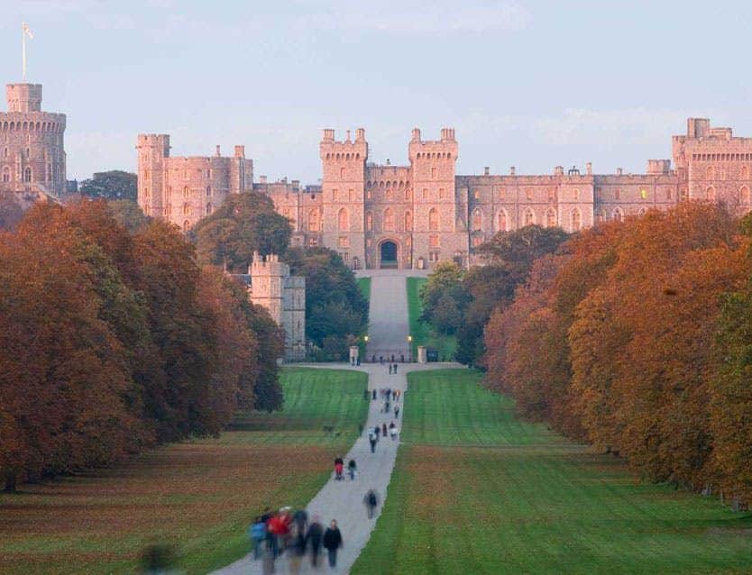 A long path leads up to a large castle with towers, surrounded by trees with autumn foliage. People walk along the path.