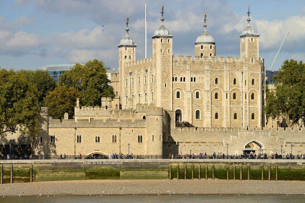 Historic stone castle with towers and battlements, seen from across a river. Group of people walking near the entrance.