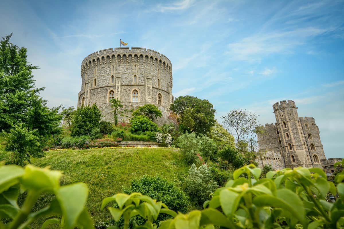 A round stone tower with battlements on top of a lush, landscaped hill adorned with various green plants and a flag flying.
