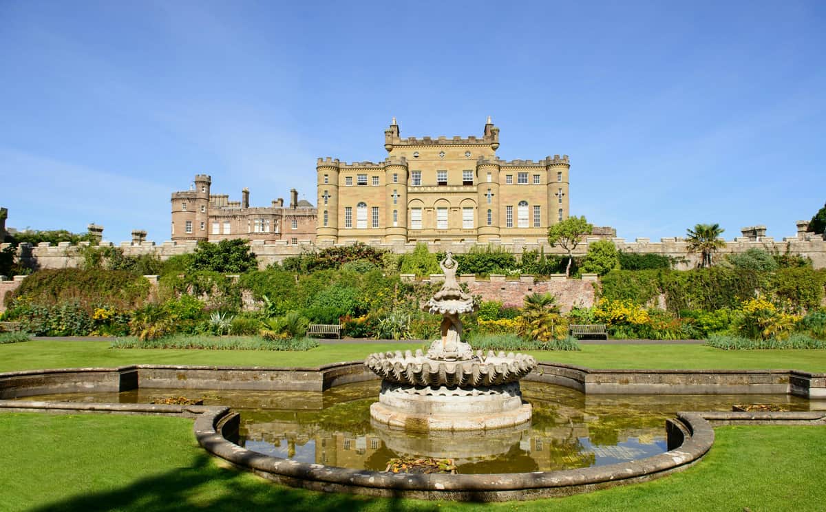 Historical castle with battlements, a manicured garden, and a tiered stone fountain in the foreground under a clear blue sky.