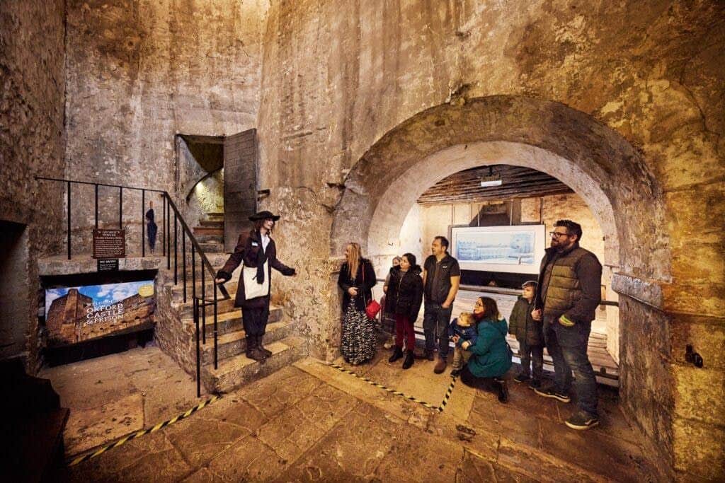 A group of people stand under a stone archway, listening to a guide inside an old, worn building. A painting is visible in the background.