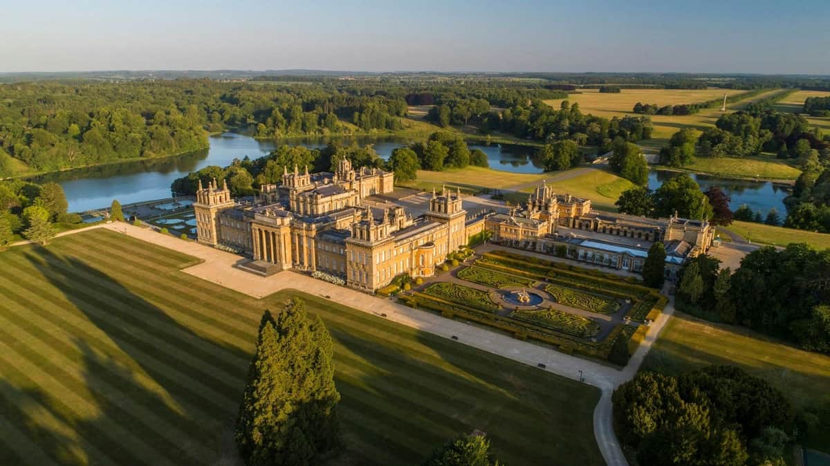 Aerial view of a large, ornate palace with extensive gardens, surrounded by trees and a lake in the distance under a clear sky.