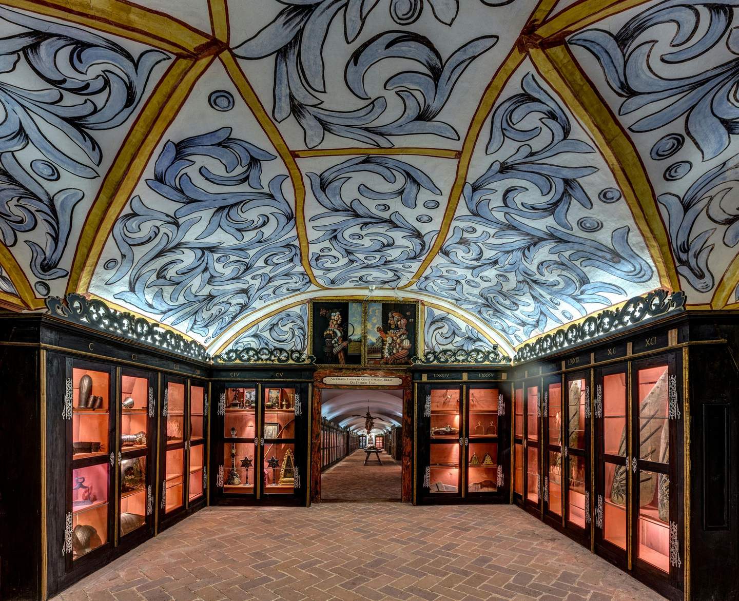 Forchtenstein Castle Ornate room with a vaulted, painted ceiling, glass display cabinets filled with artifacts, and a brick floor leading to an archway.