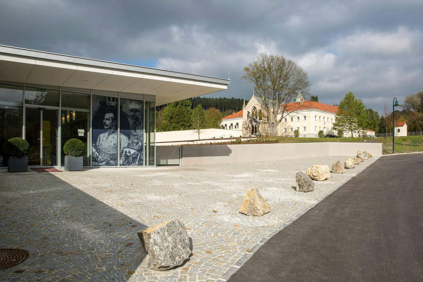 Modern building entrance with large windows and graphic art of a man in uniform, cobblestone walkway, rocks, and a historic building in the background.