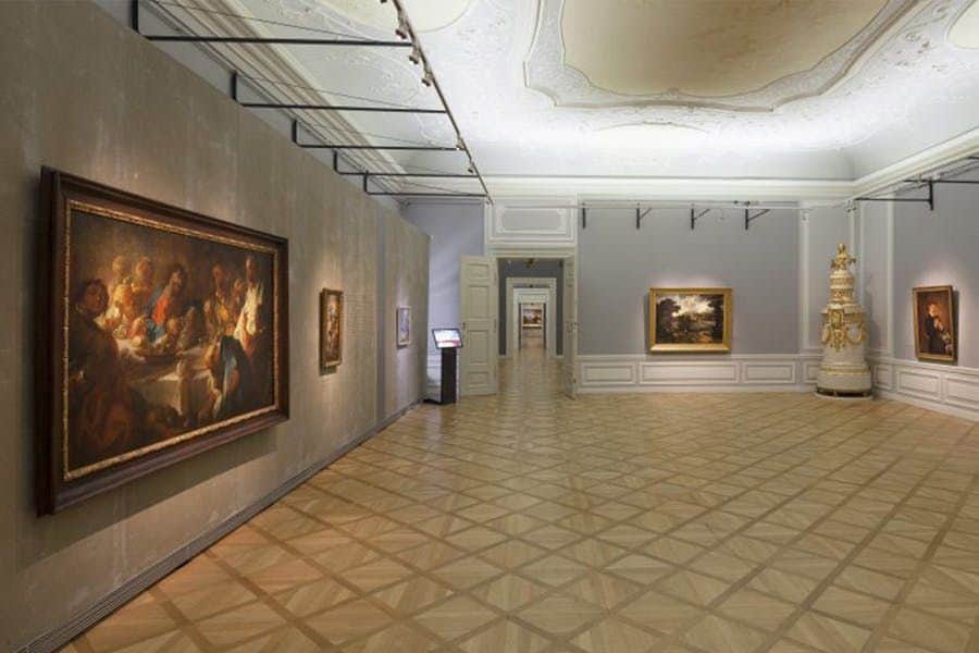 Spacious art gallery with wooden floors, framed paintings on walls, a decorative stove, and a hallway with more artwork visible.