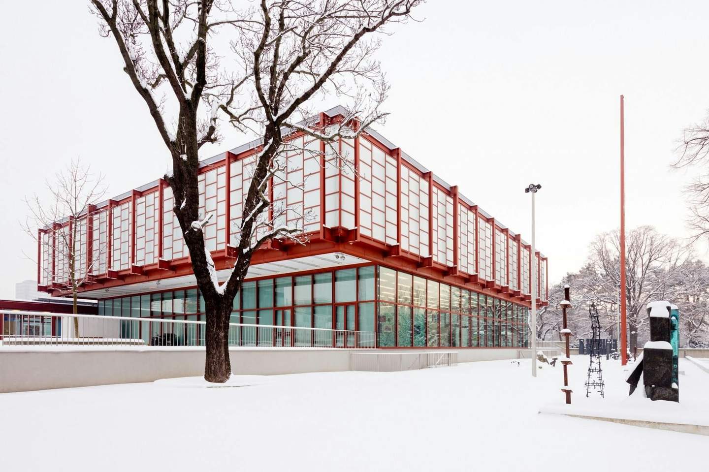A modern building with red framework and large windows in a snowy landscape, with bare trees and abstract sculptures nearby.