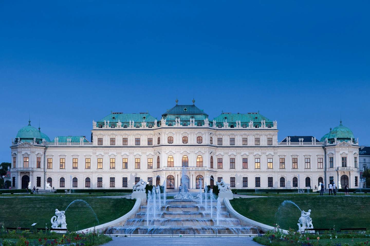 Belvedere Palace An ornate palace with green domes, fountains, statues, and gardens, set against a clear blue sky at dusk.