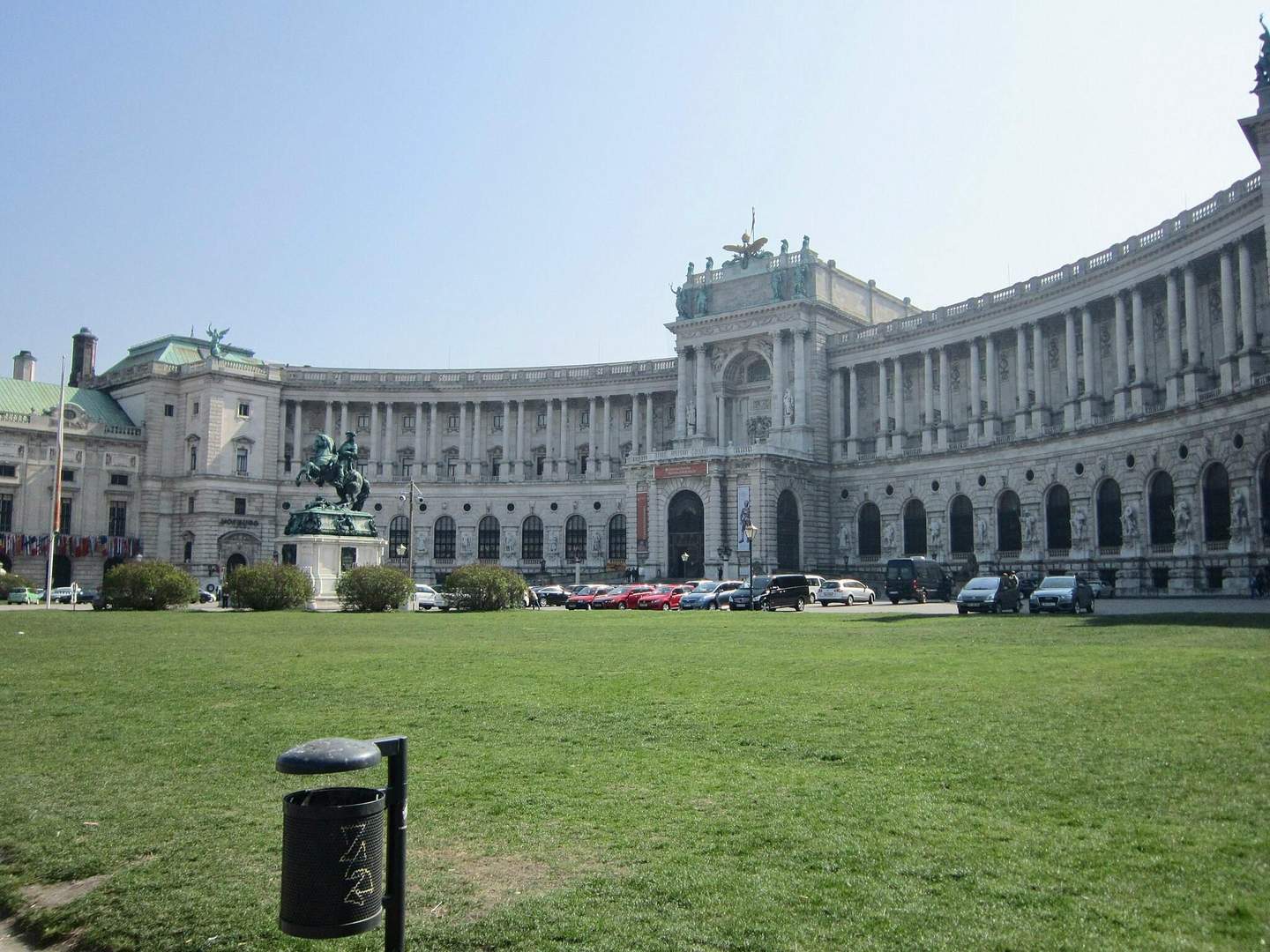 Grand building with curved facade, statue of rider on horse in front, cars parked along the street, green lawn and trash can.