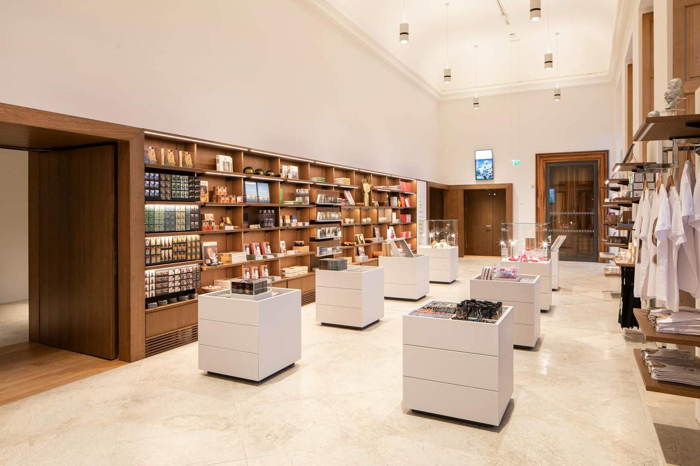 A well-lit store with shelves of books, souvenirs, and display cases. White counters and hanging t-shirts are also visible.