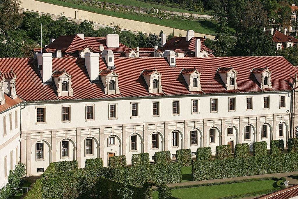 Lobkowicz Palace A long, white building with numerous windows and a red-tiled roof, surrounded by a neatly trimmed green hedge garden.