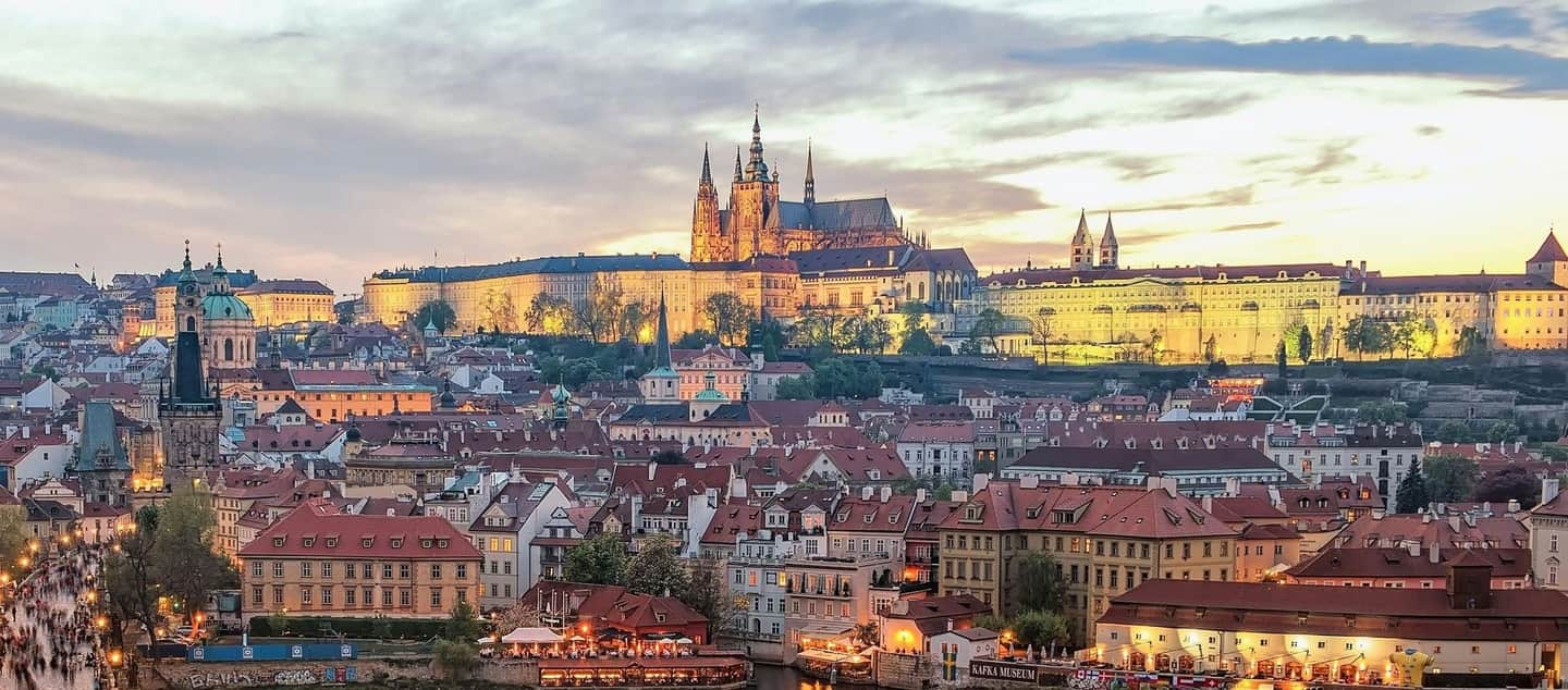 Prague Castle An evening cityscape with illuminated historic buildings, a cathedral, and red-roofed houses under a cloudy sky.